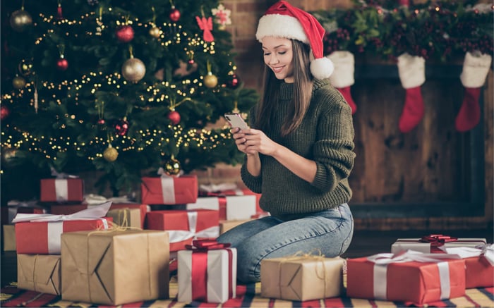 Eine Frau sitzt unter einem Weihnachtsbaum. Neben ihr liegen Geschenke. Sie schaut lächelnd auf ihr Smartphone.