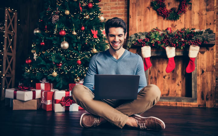 Ein Mann sitzt in einem Schneidersitz vor einem geschmückten Weihnachtsbaum und bestellt über seinen Laptop in einem Online-Shop. 