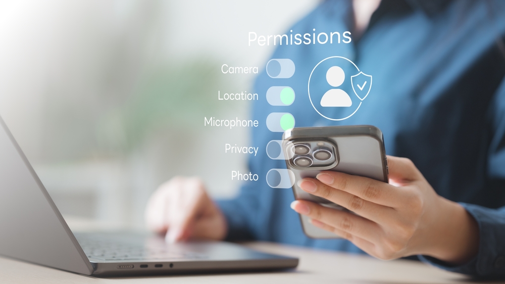 Person nutzt Smartphone neben Laptop, eingeblendete App-Berechtigungen wie Kamera, Standort und Datenschutz symbolisieren App Tracking und Privatsphäre.