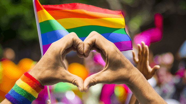 Zwei Hände bilden ein Herz vor Regenbogenfahnen.