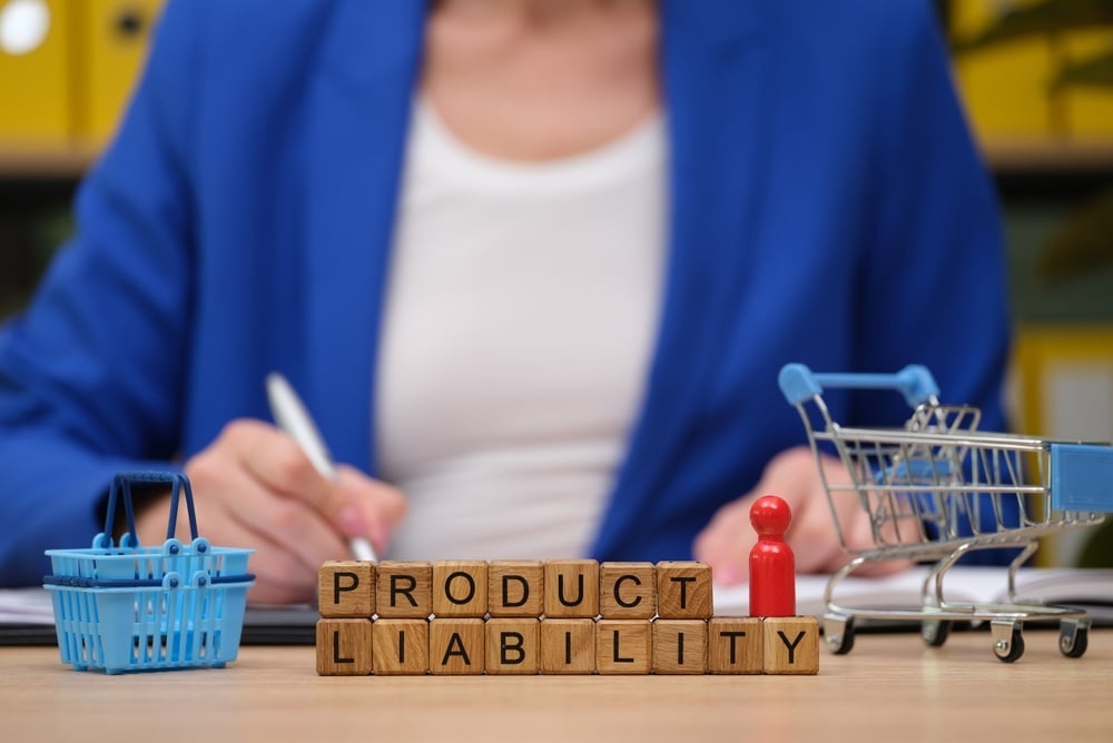 Ein Business-Profi in einem blauen Blazer schreibt Notizen an einem Schreibtisch, der mit Papierkram und einem Einkaufswagen gefüllt ist. Holzbausteine erläutern die Produkthaftung und heben die wichtigsten Diskussionspunkte hervor